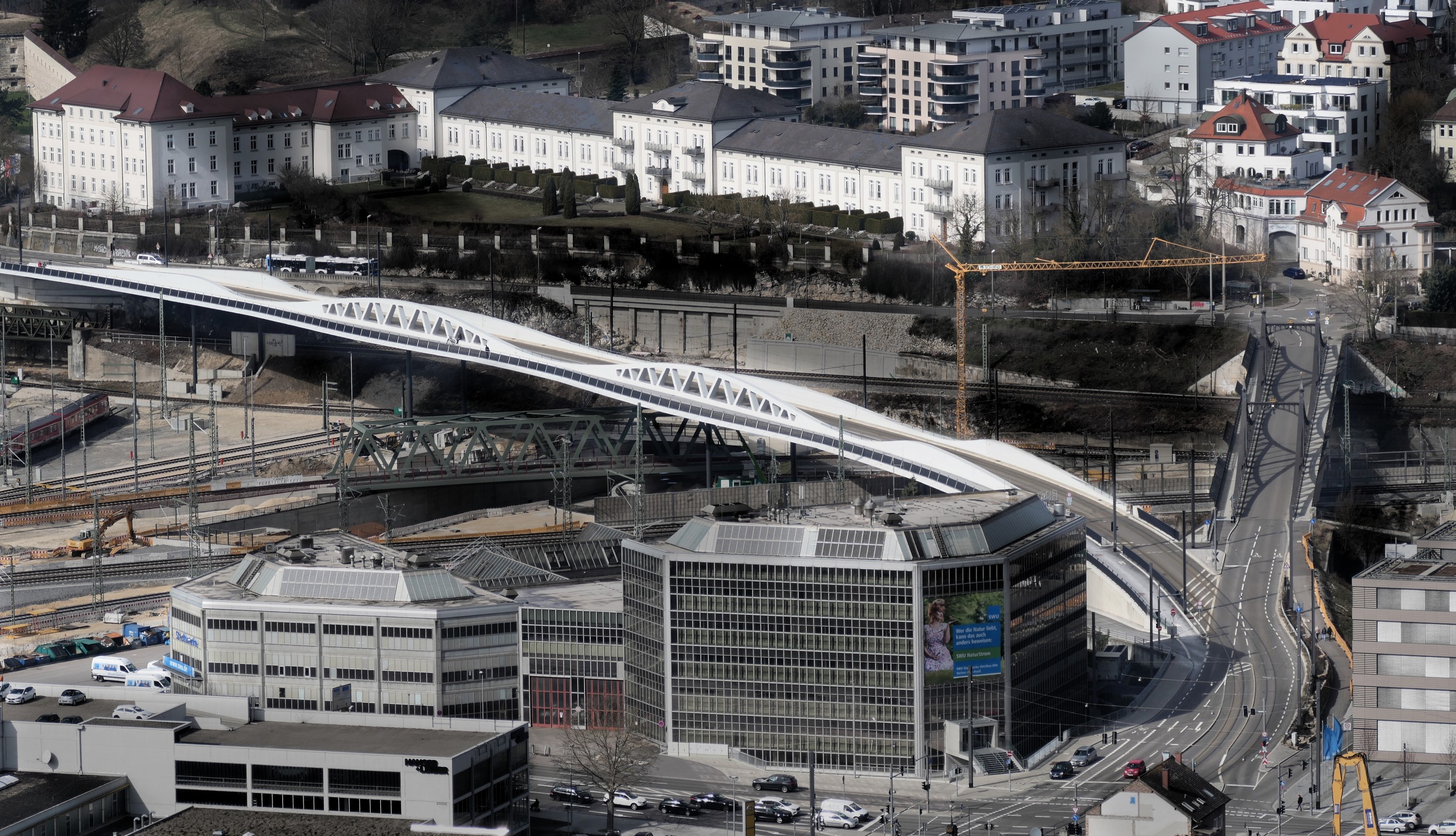 Das Bild zeigt quer verlaufend als weißes Band die neue Kienlesbergbrücke in Ulm. Sie zweigt rechts vorne hinter einem großen Gebäude ab  überspannt nach links hinten verlaufend die Gleisanlagen der Deutschen Bahn am Ulmer Hauptbahnhof.