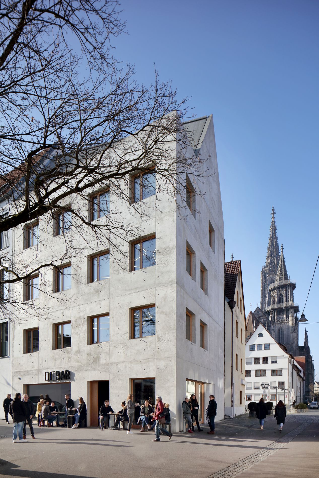 Die Abbildung zeigt von links in das Bild hineinragend das hoch aufragende, fünfgeschossige Wohn- und Geschäftshaus k5 in der Altstadt von Ulm. Es hat eine glatte Betonfassade, quadratische Fenster und ein Satteldach. Im Erdgeschoss befindet sich ein Café, draußen sind Personen an kleinen Tischen und Passanten. Rechts im Hintergrund sind die Türme des Ulmer Münsters zu sehen.
