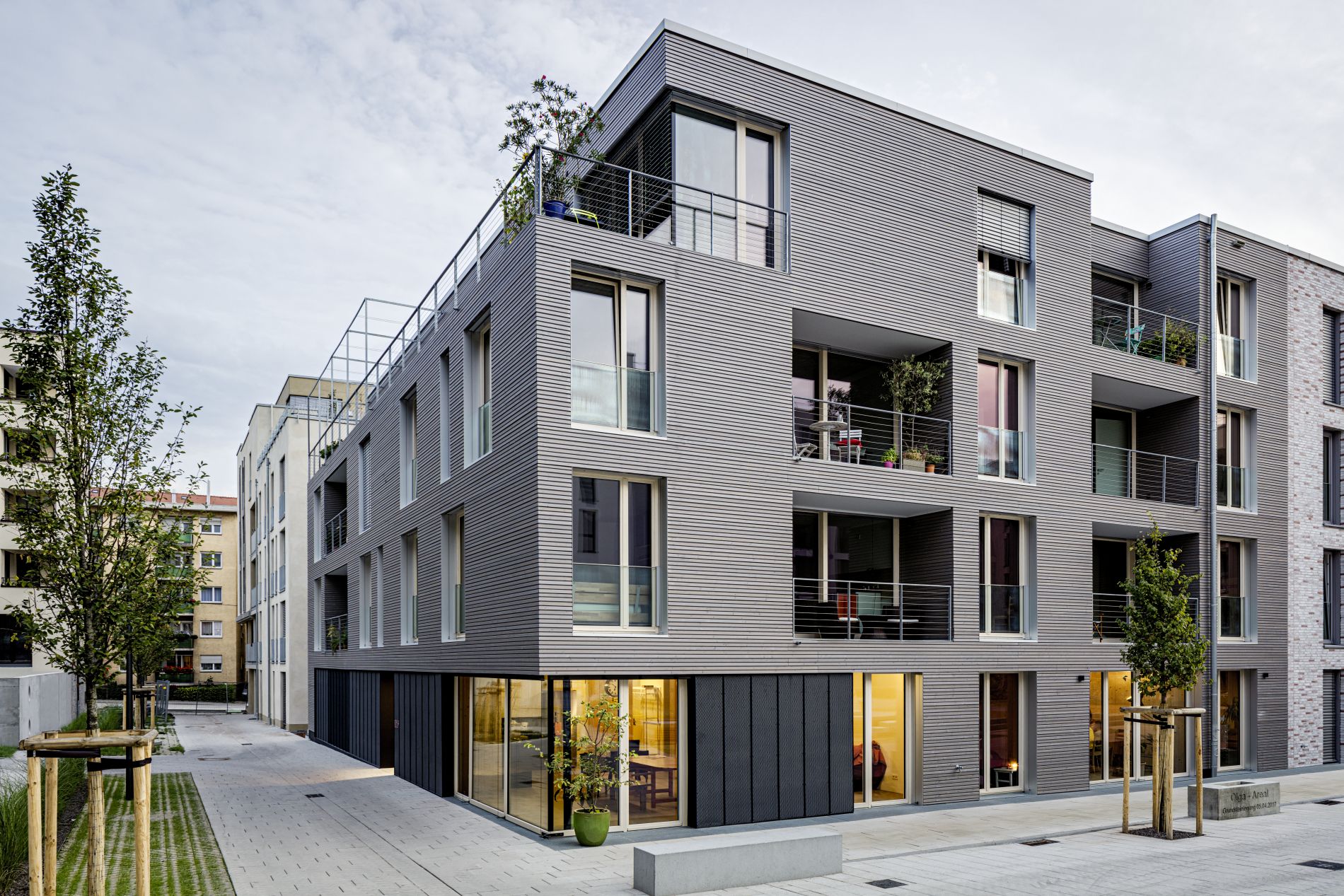 Im Bildvordergrund ist das viergeschossige Wohnhaus namens "MaxAcht" in Stuttgart zu sehen, dessen Fassade große Fenster und Loggien hat und mit querverlaufenden Holzbrettern verkleidet ist.