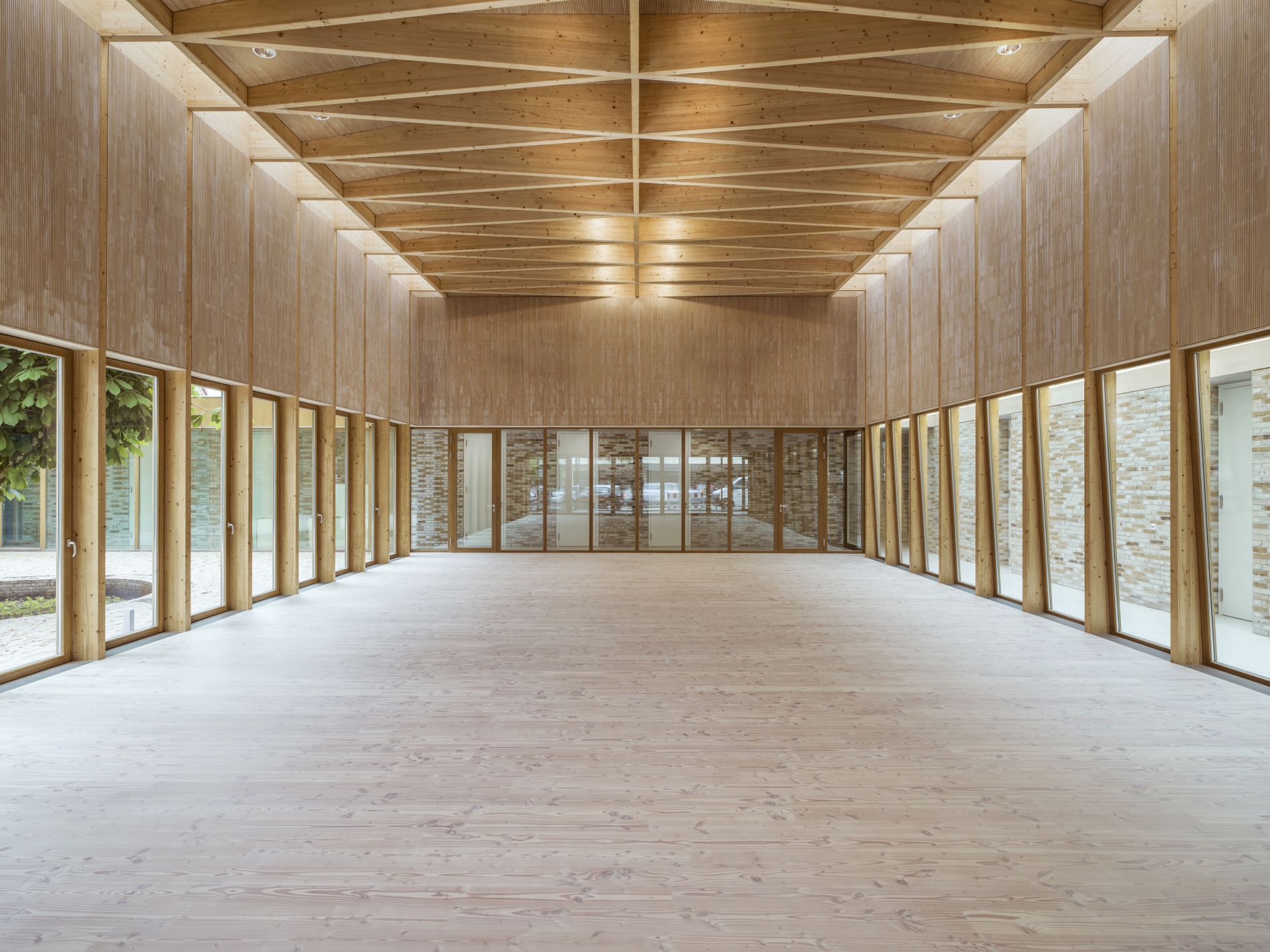 Der Blick ist in die Tiefe eines langgestreckten Saals mit einem hellen Holzfußboden gerichtet. Die seitlichen Wände haben hohe Fenster bzw. Glastüren, von denen die auf der rechten Seite nach innen geneigt sind. Ein kreuzweise verlegtes Holztragwerk überspannt den Saal stützenfrei.