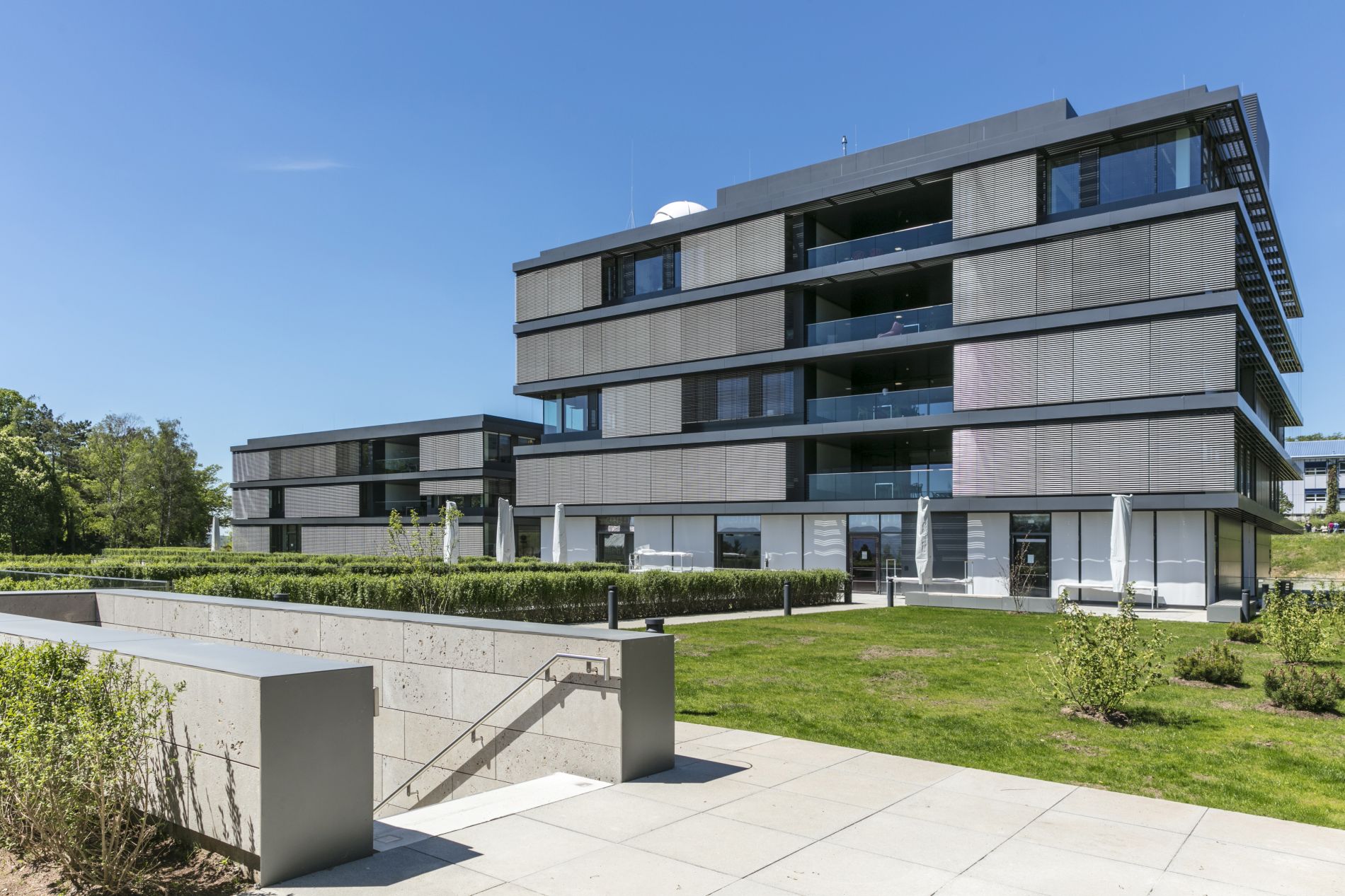 Das Bild zeigt die Ost-Ansicht des Max-Planck-Instituts für Intelligente Systeme in Tübingen: Dies sind zwei Gebäude mit Fassaden aus großen Glasflächen und Gesimsen aus Aluminium, von denen das rechts stehende mit fünf und das linke, weiter hinten stehende mit drei Geschossen zu sehen sind.