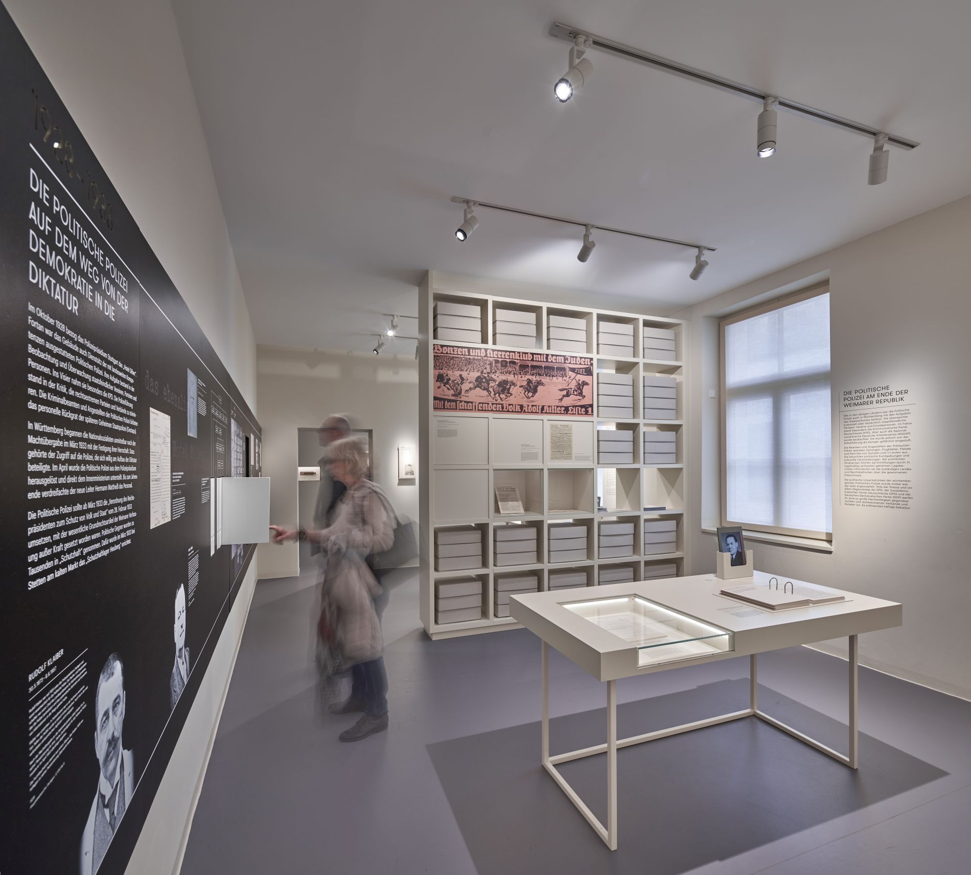 Das Bild zeigt den Blick in einen Ausstellungsraum im Lern- und Gedenkort "Hotel Silber" in Stuttgart. An der links nach hinten verlaufenden Wand des weiß gehaltenen Raums mit hellgrauem Boden befindet sich eine schwarze Informationstafel mit weißer Schrift. In der Bildmitte befindet sich, den Raum in der Tiefe teilend, ein raumhohes weißes Regal, in dem weiße Kartons liegen, die an Akten erinnern sollen.