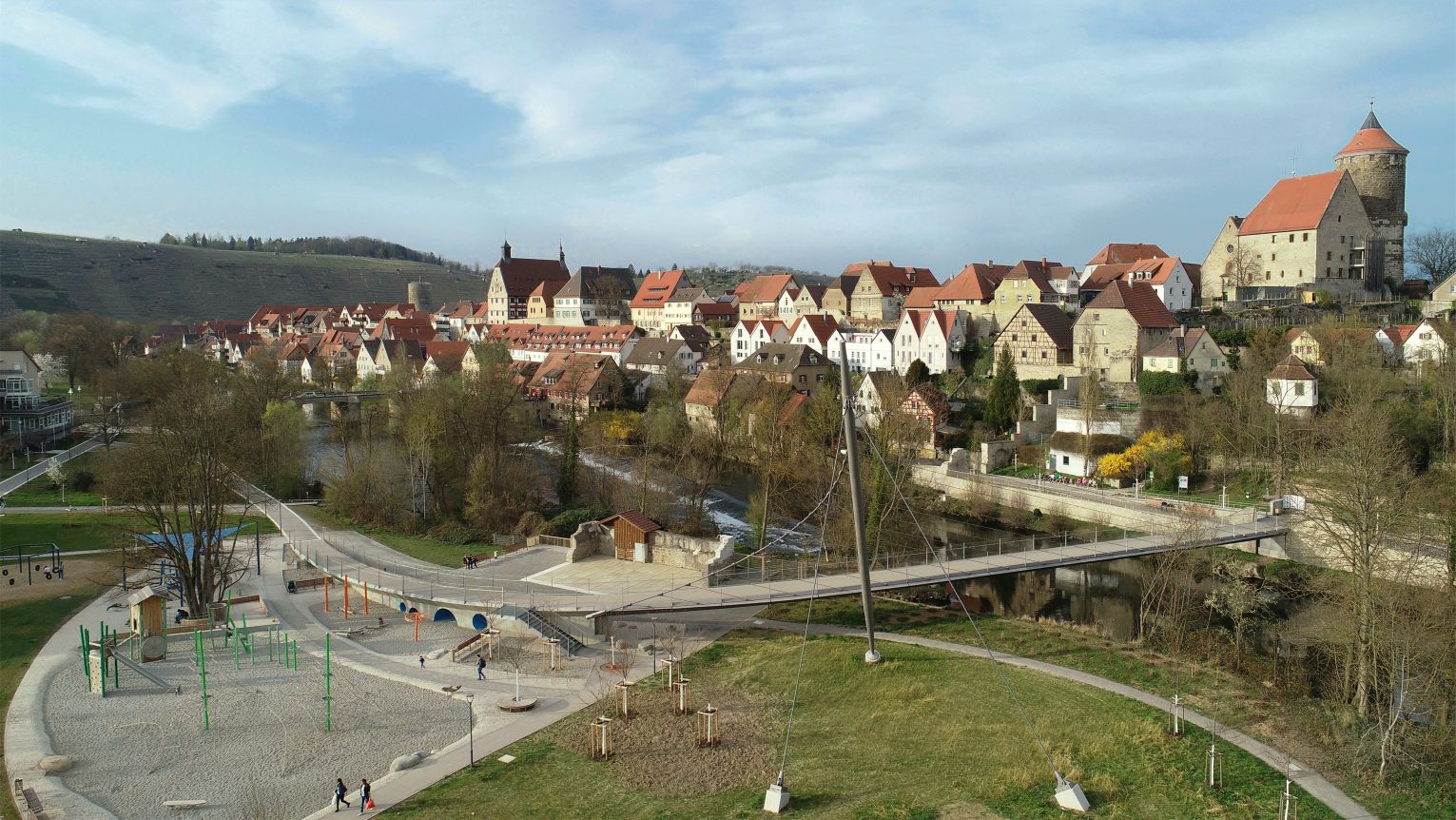 Die mit seilgespannte Fußgängerbrücke und Stahlbetonrampen verbindet die beiden Stadtteile im Hintergrund und links im Bild zu sehen und gibt gleichzeitig den Blick frei auf die Enzaue rechts im Bild. 