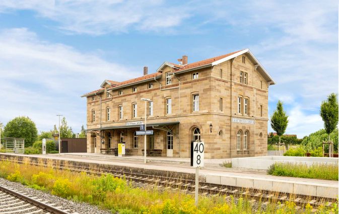 Die markante Natursteinfassade eines denkmalgerecht sanierten Bahnhofsgebäudes im Hintgerund, direkt an den im Vordergrund abgebildeten Gleisen.