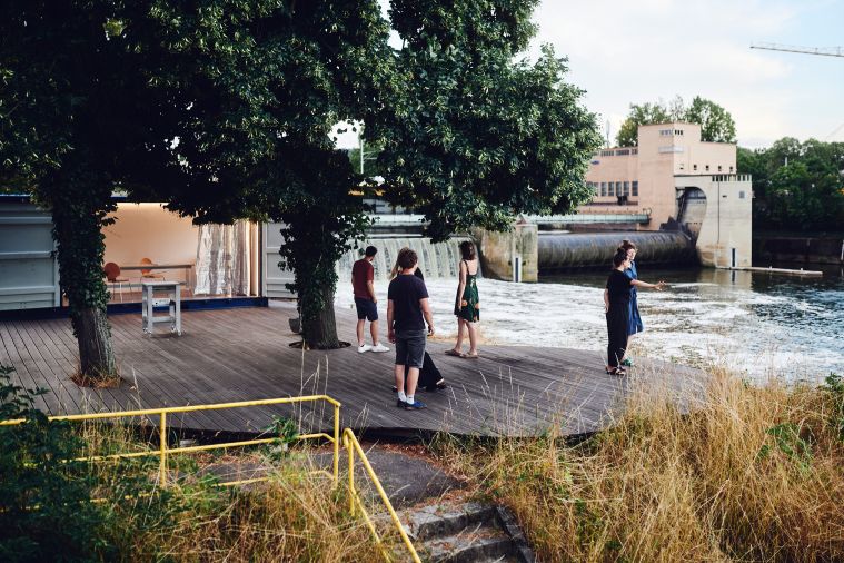 Die Terrasse und Liegewiese der Neckarinsel mit umgebautem Überseecontainer umgeben von Schleuse und Wasserkraftwerk, im hinteren Teil des Bildes zu sehen. 