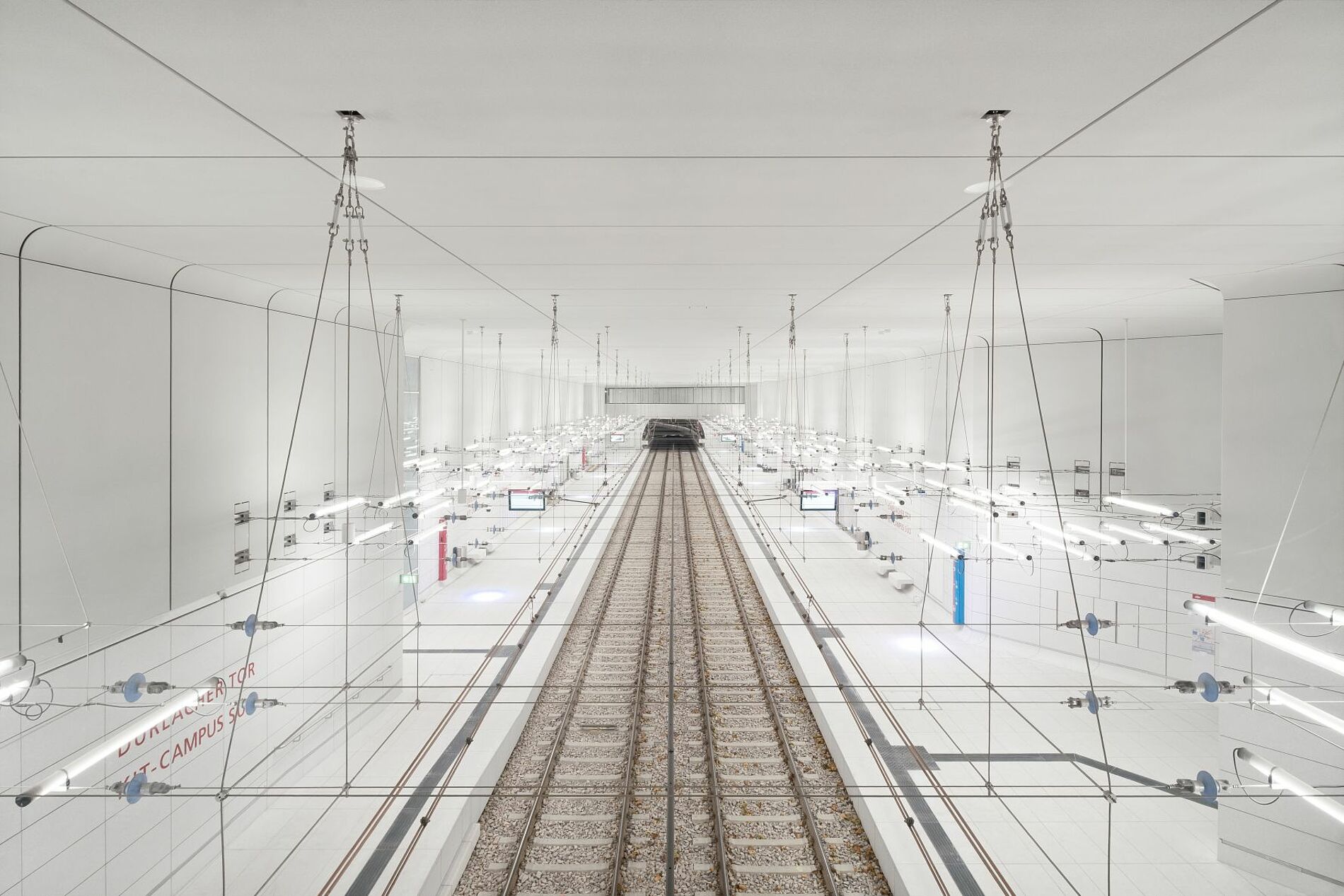 Unterirdische Stadtbahn Karlsruhe mit weißen Raumschalen auf Bahnsteigebene und abgehängter Stahlseilkonstruktion mit integriertem Lichtkonzept.